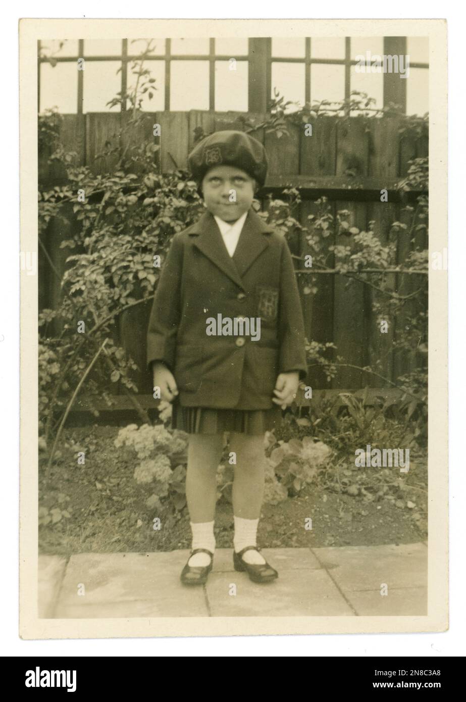 Originelles, amüsantes Foto eines jungen Mädchens, das ein lustiges Gesicht zieht, Schuluniform trägt, mit Jacke und Baskenmütze, für ihren ersten Tag auf dem Souvenirfoto der Grundschule posiert, um 1930er oder 1940er, Großbritannien Stockfoto