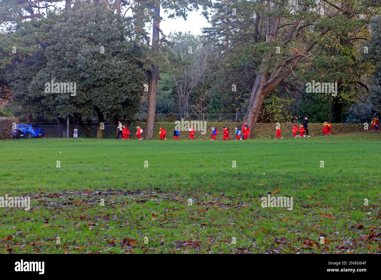 Kindergärtnerinnen in einer Reihe mit roter Kleidung folgen Lehrern durch den Park in Sophia Gardens, Cardiff, 2022. Verschwommene Kinder, unidentifizierbar. Stockfoto