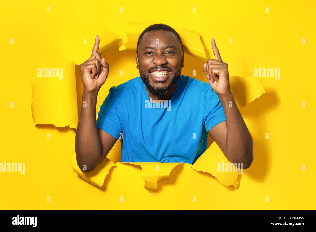 Porträt eines aufgeregten afroamerikanischen Mannes, der mit den Fingern nach oben zeigt und lächelt, der in einem zerrissenen Papierloch auf gelbem Hintergrund steht Stockfoto
