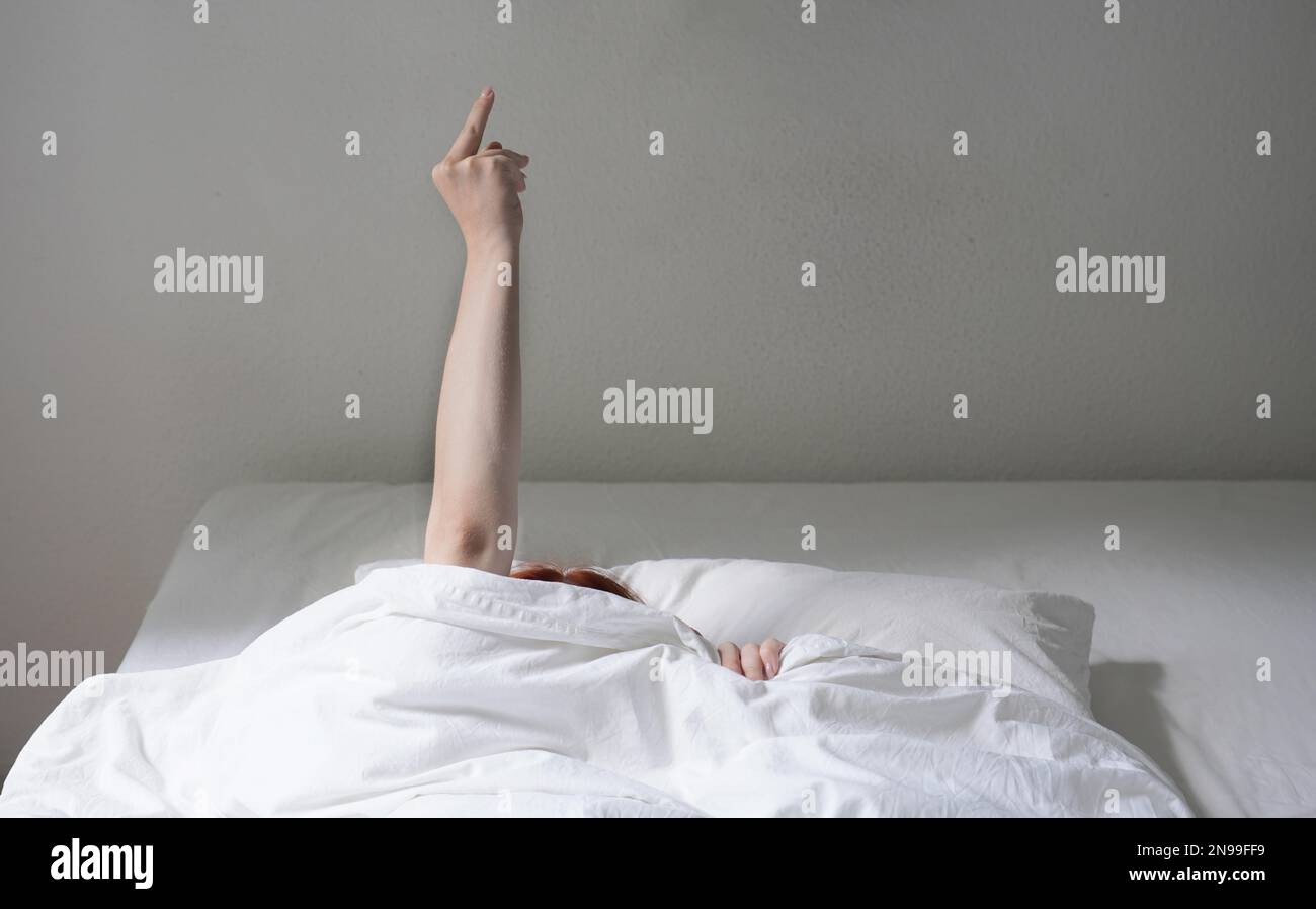 Frau, die den Finger umdrehte, während sie sich unter der Decke versteckte und sich weigerte, aus dem Bett zu gehen Stockfoto