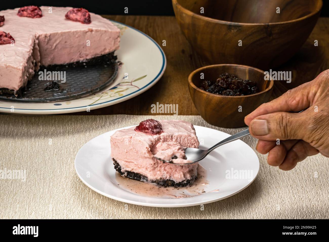 Männlicher Senior Hand mit Metalllöffel, der einen Bissen von köstlichem hausgemachten gefrorenen Erdbeerkäsekuchen in weißer Keramikschale auf einer Tischmatte mit getrocknetem Stockfoto