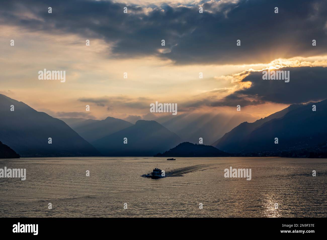 Am späten Abend im Sommer am comer See mit trüben Bergen, Sonnenbalken, die durch Wolken scheinen, und Tragflächenboot, das sich bellagio nähert Stockfoto