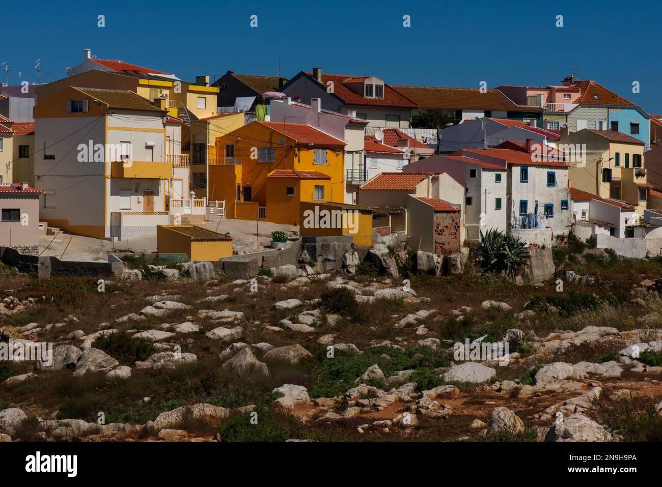 Leuchtendes Orange inmitten von Gelb-, Blau-, Rosa- und Weißtönen: Moderne Häuser und Apartments auf Klippen, die auf felsigen Fundamenten über dem Hafen von Peniche im Stadtteil Leiria an der Atlantikküste von West-Zentral-Portugal erbaut wurden. Stockfoto