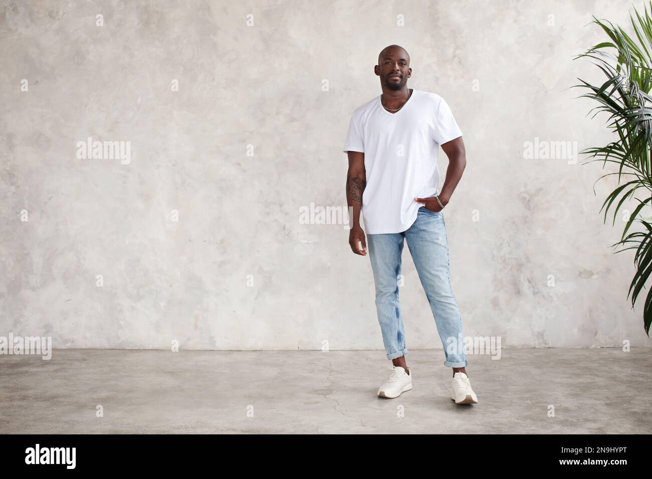 Ein hübscher junger afroamerikanischer Mann steht mit vollem Körper, schaut in die Kamera, gegen die hell strukturierte Wand im Studio. Männliches Model trägt lässige Kleidung in Weiß Stockfoto