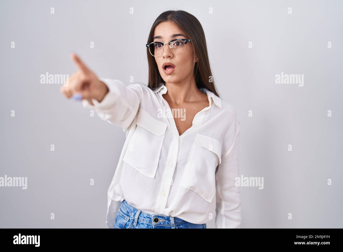 Junge brünette Frau mit Brille, die mit dem Finger nach vorne zeigt, mit offenem Mund überraschter Ausdruck, etwas auf der Vorderseite Stockfoto