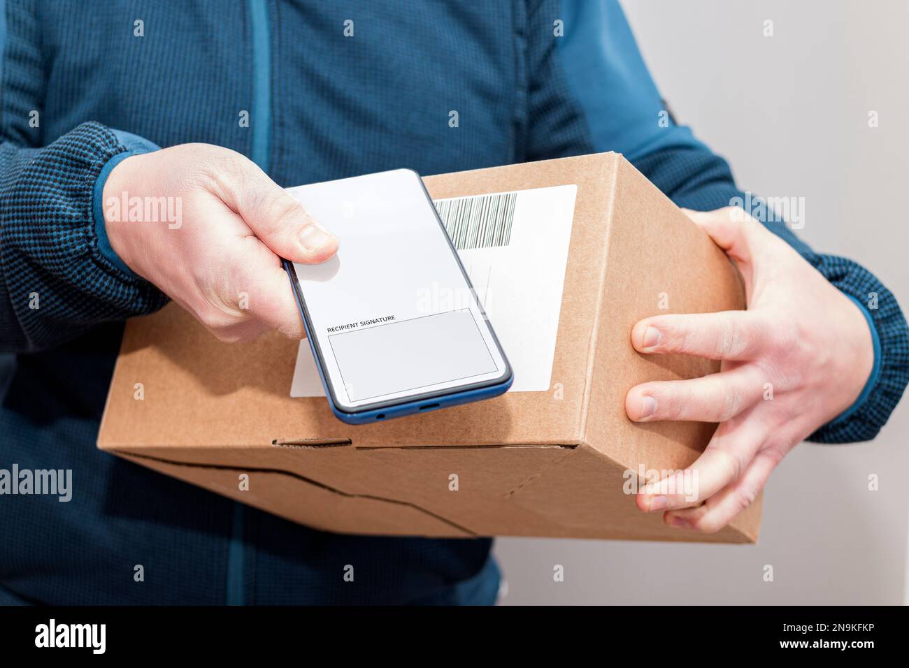 Nahaufnahme der Hände eines unbekannten Zustellers, der ein Paket mit einem Mobiltelefon in der Hand hält und einen Unterschriftsbildschirm anzeigt. Stockfoto