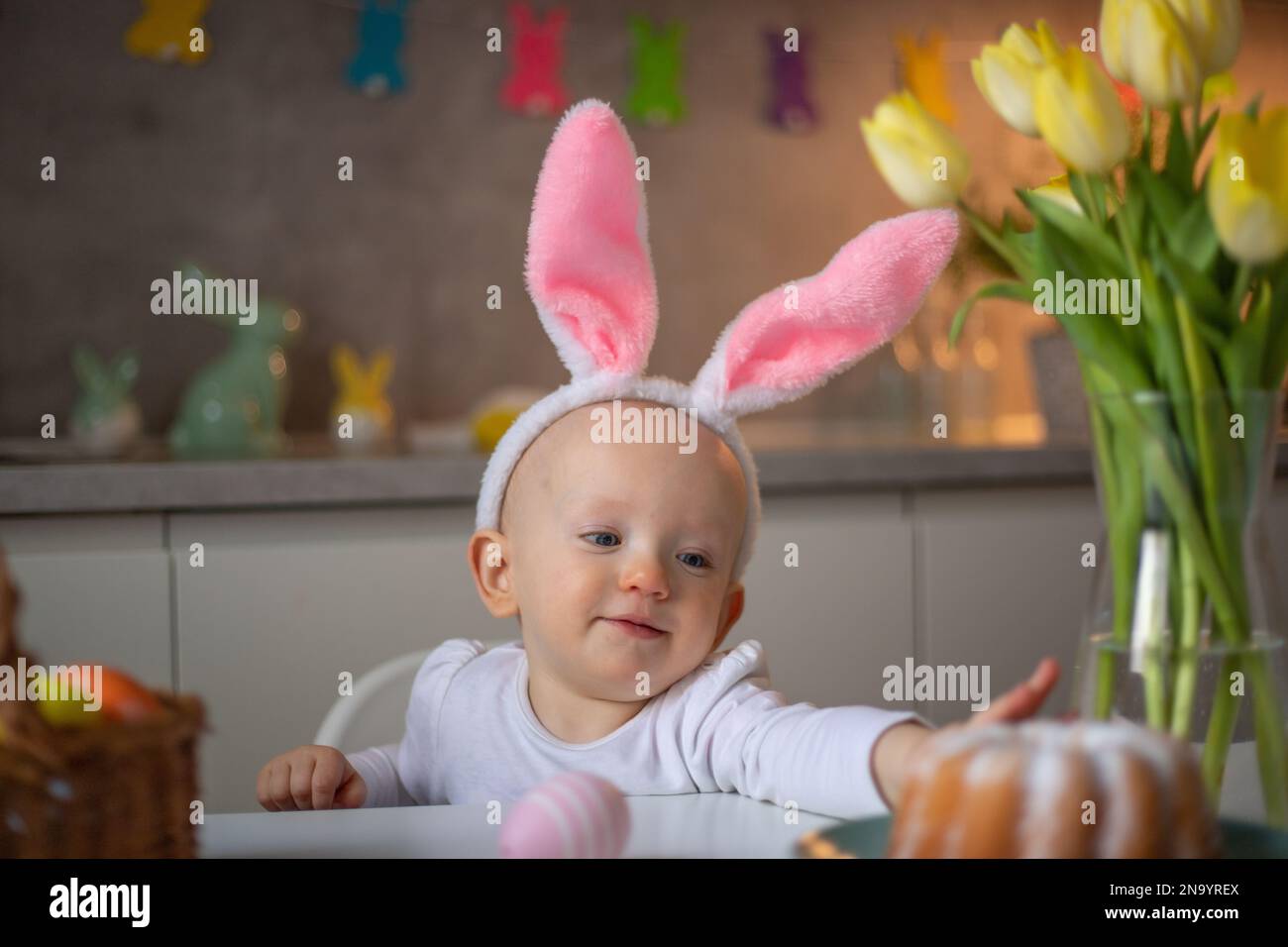 Fröhliches süßes kleines Mädchen mit Hasenohren an ostern, das am Tisch in der Küche sitzt. Stockfoto