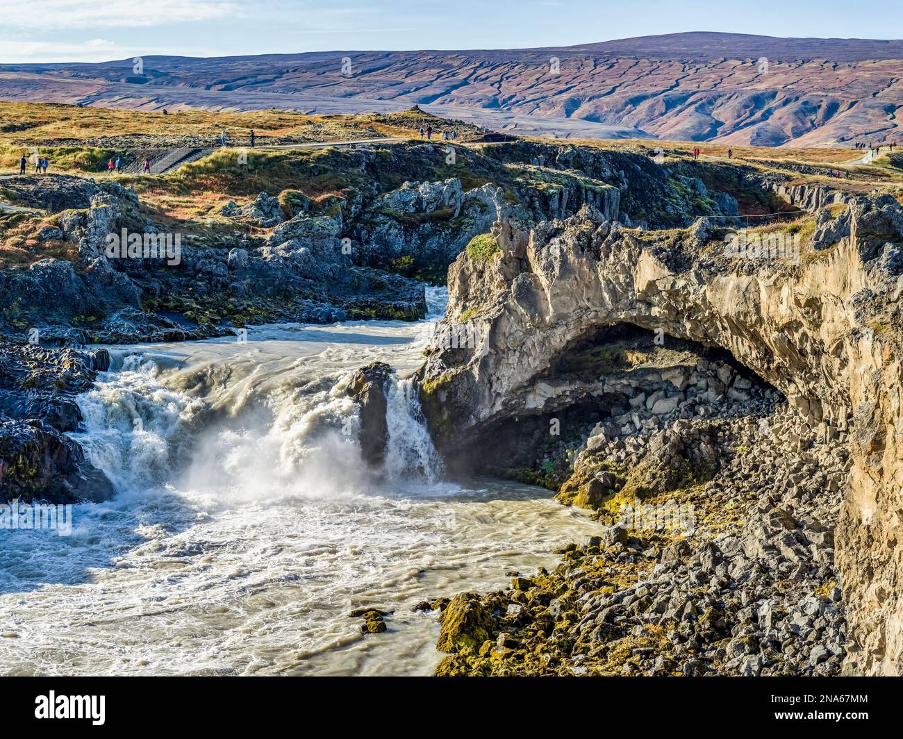 Wasserfall und Fluss fließen durch zerklüftetes Gelände mit Touristen auf einem Wanderweg; Thingeyjarsveit, Nordostregion, Island Stockfoto