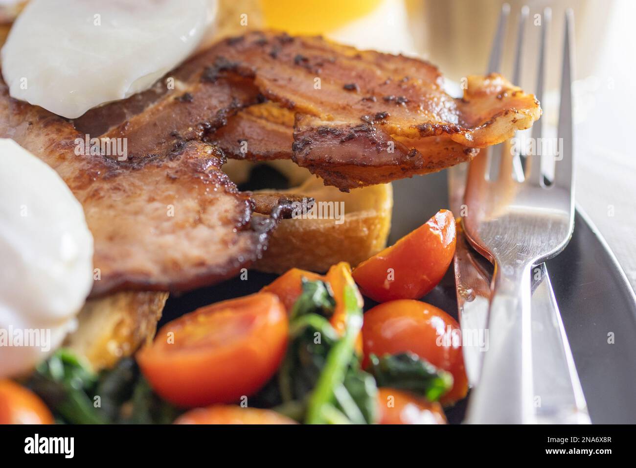 Großes Frühstück, pochierte Eier mit Speck auf Toast mit gebratenen Tomaten und Spinat und einem Getränk aus Orangensaft, schwarzer Platte mit Messer und Gabel Stockfoto