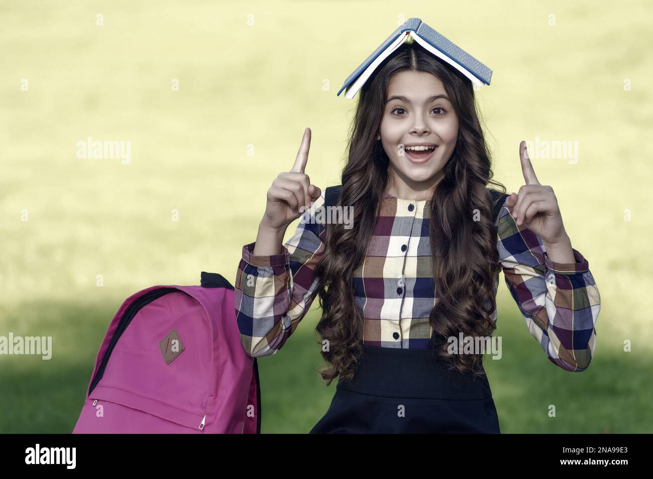 Happy kleines Kind in der Schule Uniform halten Buch auf dem Kopf zeigen Zeigefinger nach oben sonnig im Freien, Idee Stockfoto