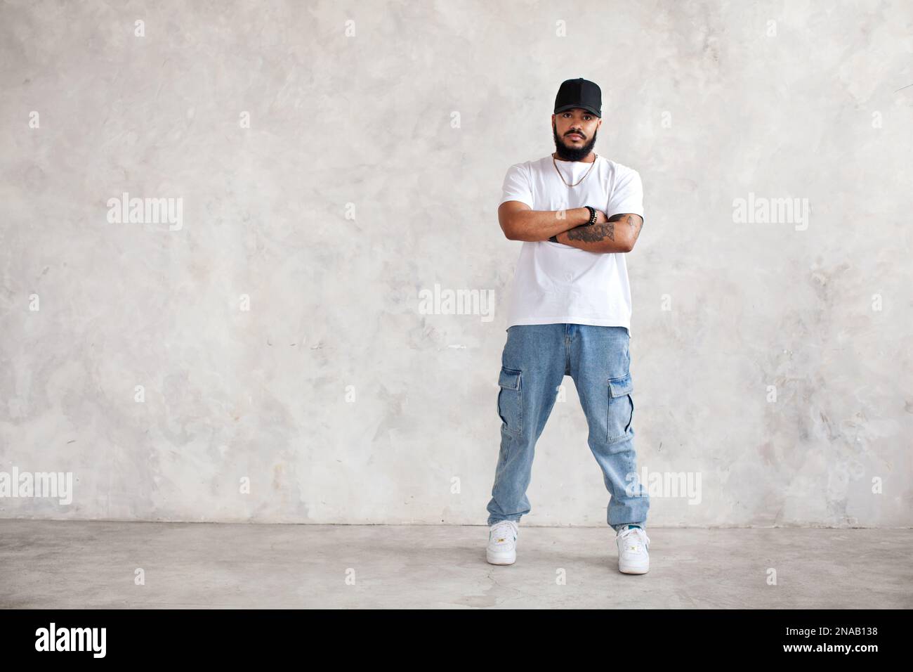 Ein ganzer Porträt Hispanic Mann steht die Arme gekreuzt, in lässigen Jeans und weißem Modell-T-Shirt und Baseballmütze, der coole Typ schaut auf die Kamera Agains Stockfoto