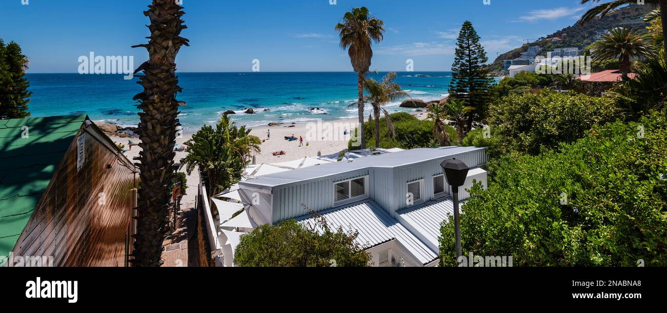 Sonnenanbeter genießen den weißen Sandstrand und das blaue Meer mit Häusern entlang der Küste, Südafrika Stockfoto