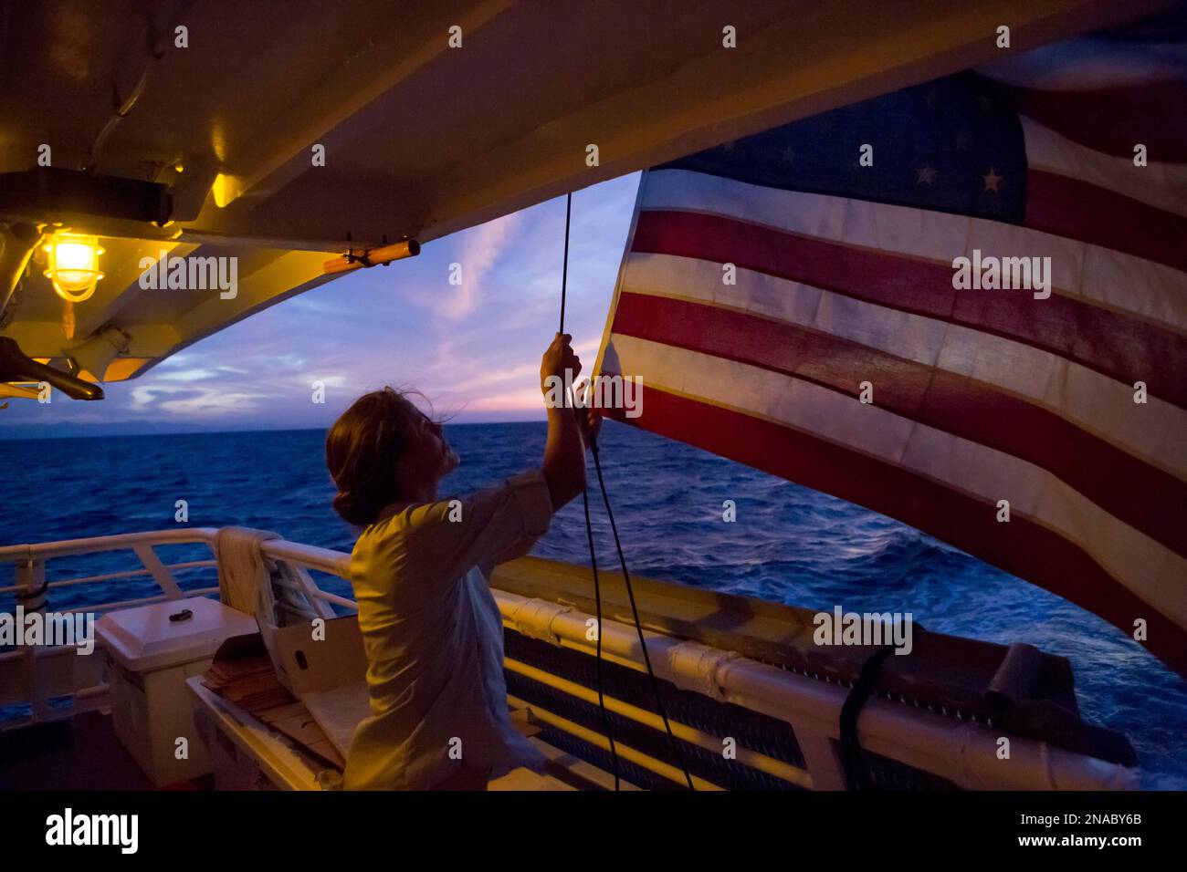 Inmitten eines farbenfrohen Morgenhimmels in der Nähe von Panama hebt ein Deckblatt an Bord eines Expeditionsschiffs bei Sonnenaufgang im Coiba-Nationalpark, Panama, die amerikanische Flagge Stockfoto