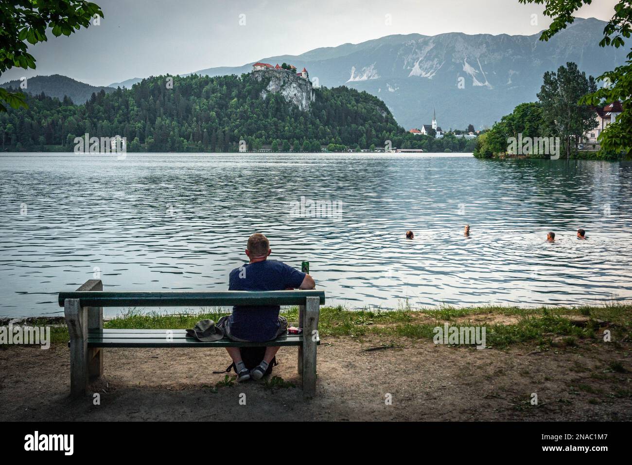 Rückansicht eines allein entspannten Mannes, der auf einer Bank neben einem Bleder See sitzt, mit einer Dose Bier in der Hand, die schwimmenden Menschen und das Schloss Bled betrachtet Stockfoto