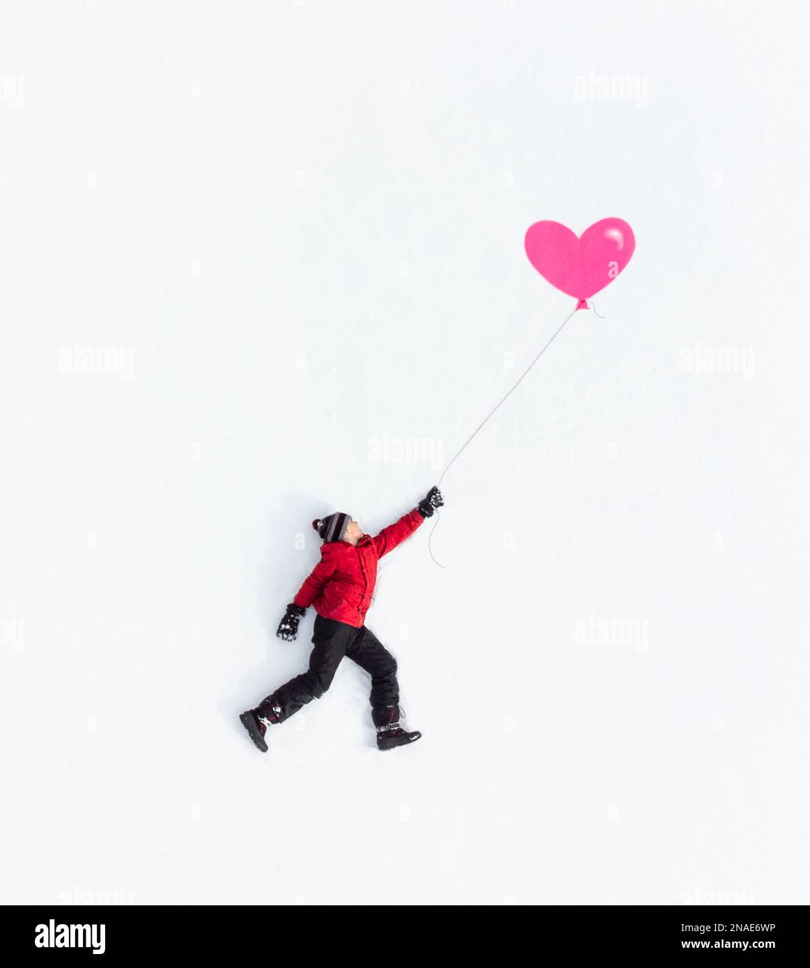 Luftaufnahme eines Jungen, der in Winterkleidung mit pinkem Ballon auf Schnee liegt. Stockfoto