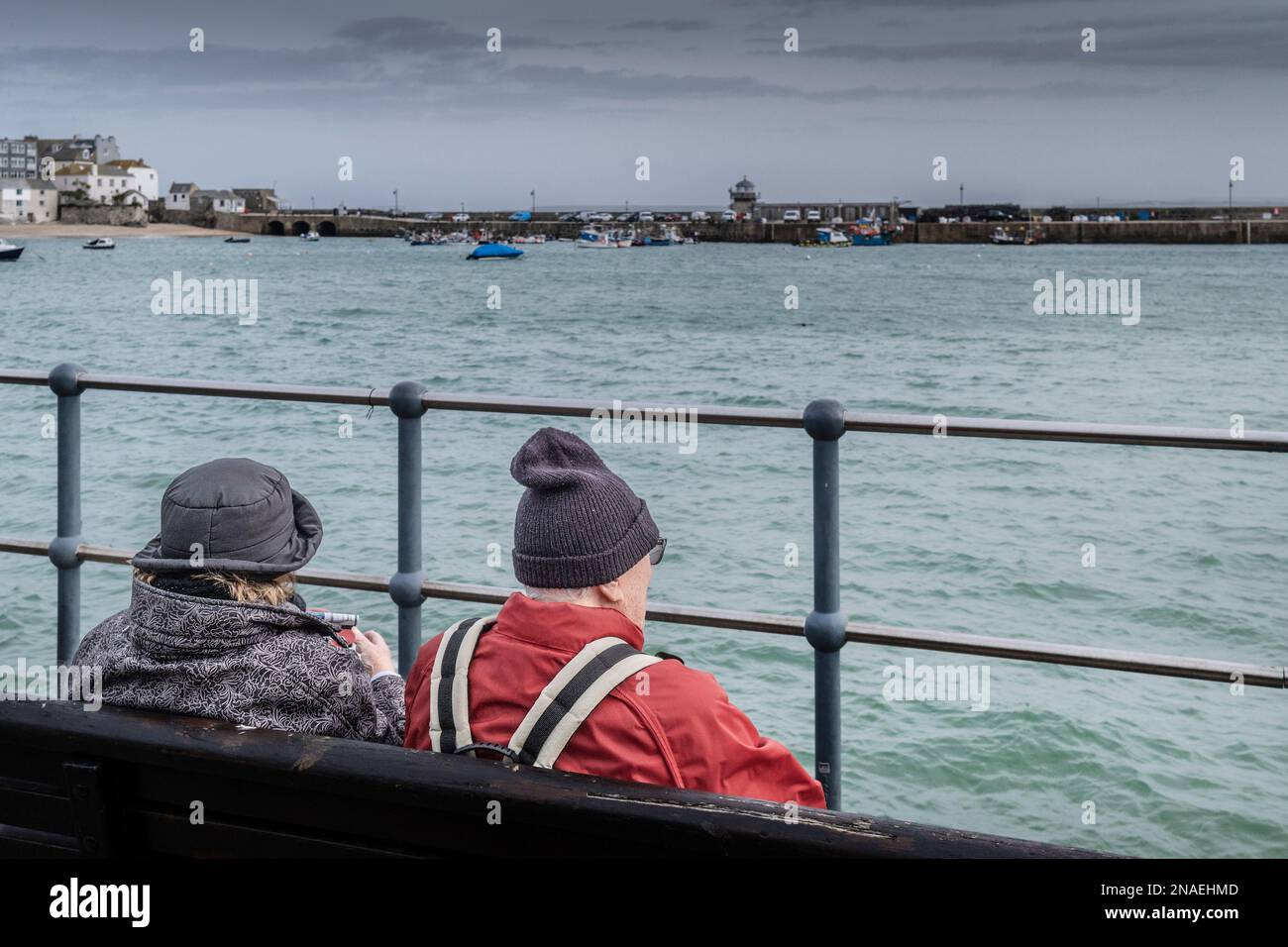 Wetter in Großbritannien. Besucher sitzen an einem regnerisch kalten, elenden Tag in der historischen Küstenstadt St. Ives in Cornwa auf einer Bank mit Blick auf den Smeatons Pier Stockfoto