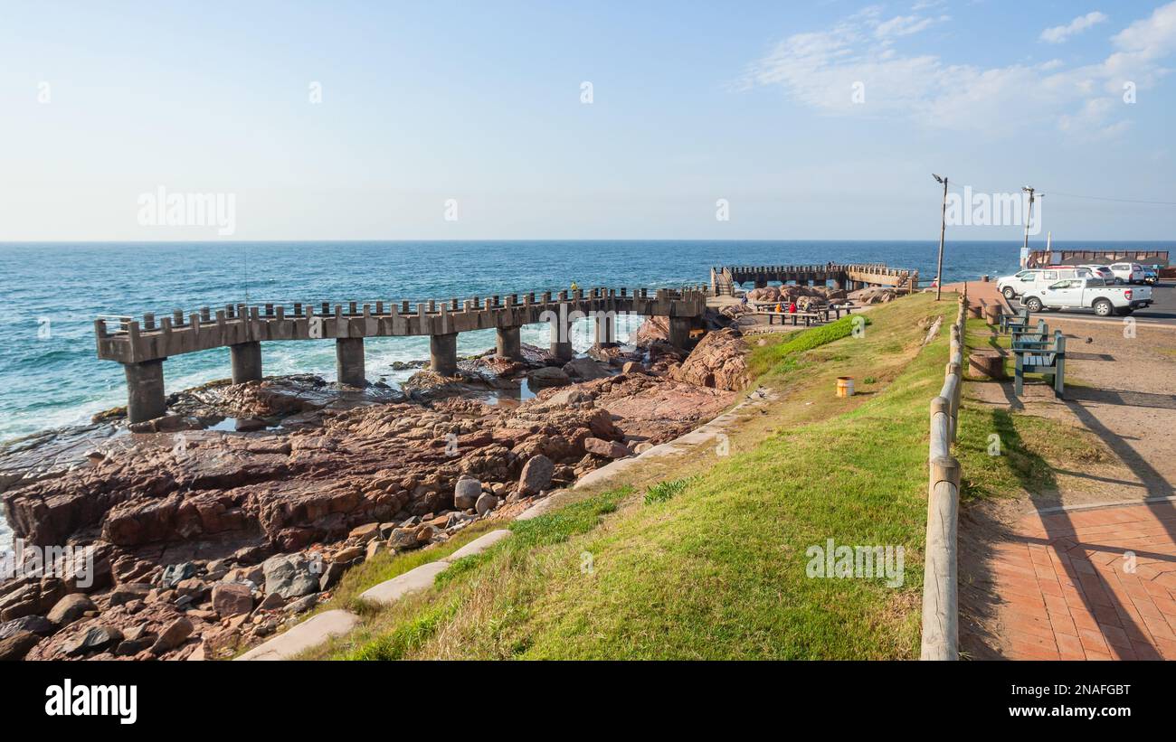 Angelpier Betonsteg an der felsigen Küste mit Blick auf das malerische blaue Meer am Sommertag mit Fischerfahrzeugen auf dem Parkplatz. Stockfoto