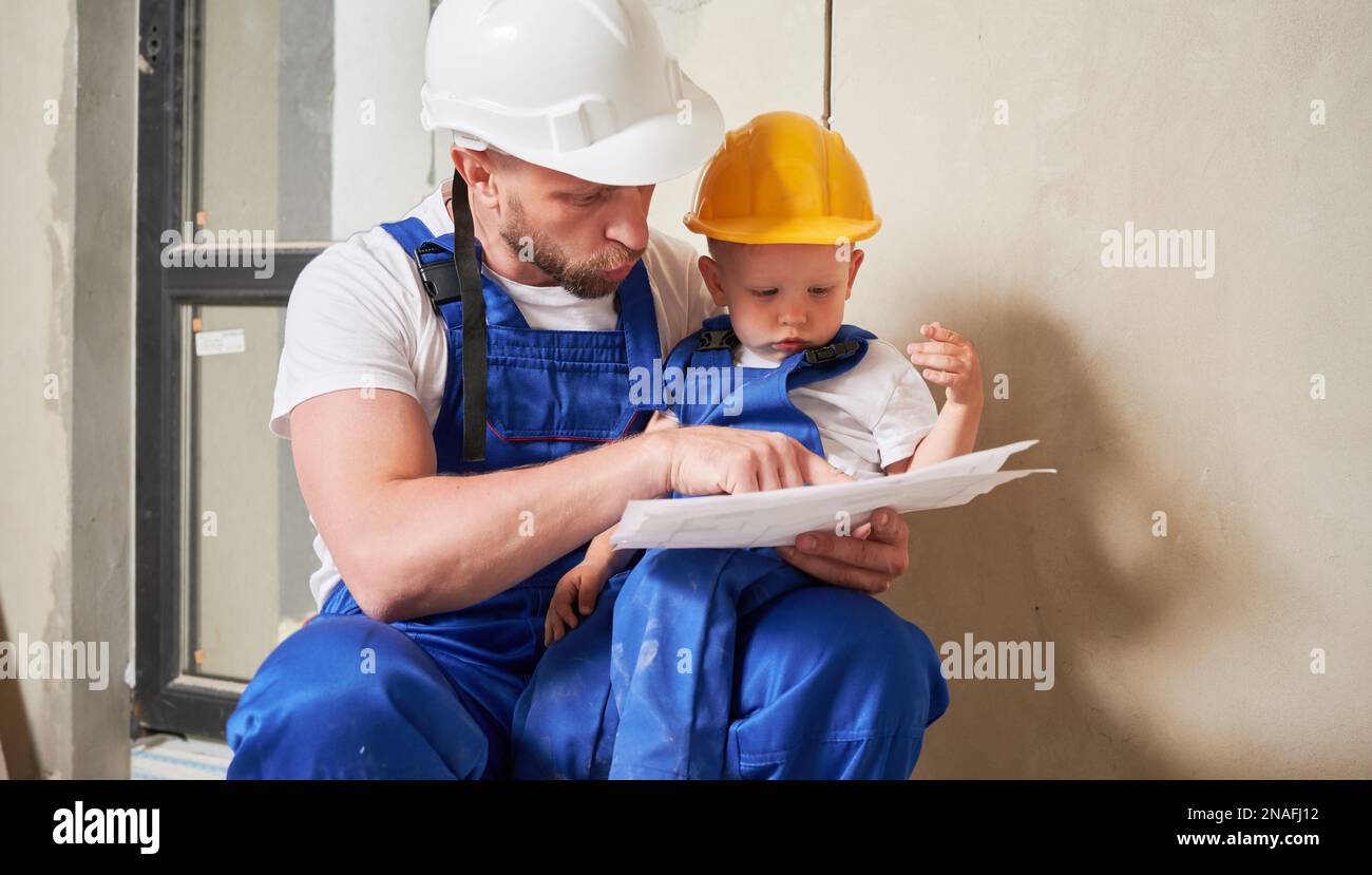 Der Baumeister zeigt dem kleinen Jungen Architekturzeichnungen. Ein bezauberndes Kind im Bausicherheitshelm, das auf dem Schoß des Vaters sitzt und den Bauplan in einer Wohnung studiert, die gerade renoviert wird. Stockfoto