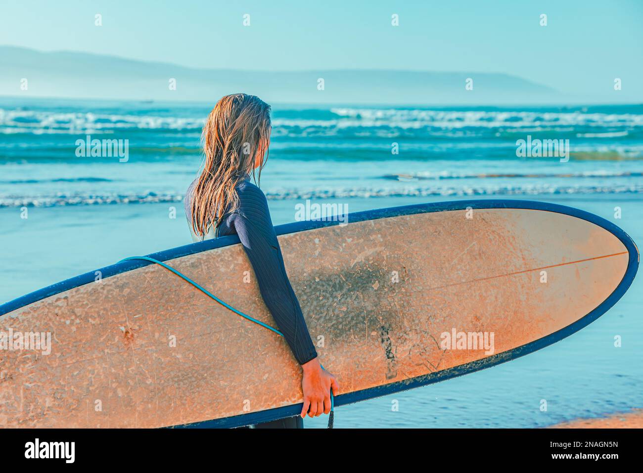 Eine weibliche Strandgängerin steht am Ufer mit einem Surfbrett in der Hand und blickt auf den Horizont des Ozeans hinaus Stockfoto