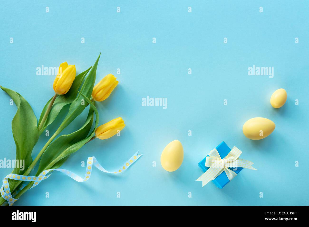 Osterfest-Hintergrund. Gelb bemalte Eier, Geschenkbox und Tulpen auf blauem Tisch. Draufsicht, flach liegend, Kopierbereich. Stockfoto