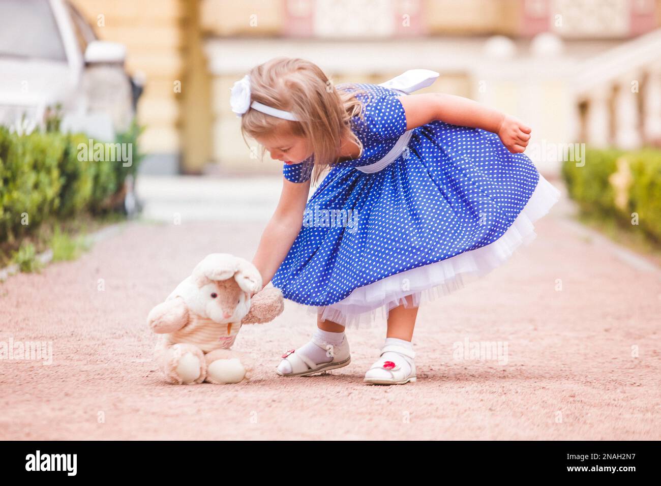 Das kleine Mädchen auf der Straße, das mit einem Spielzeug spielt. Baby legt Spielzeughase auf den Boden. Ein Mädchen in einem üppigen blauen Kleid, das in der Stadt Kiew (Ukraine) spaziert. Stockfoto