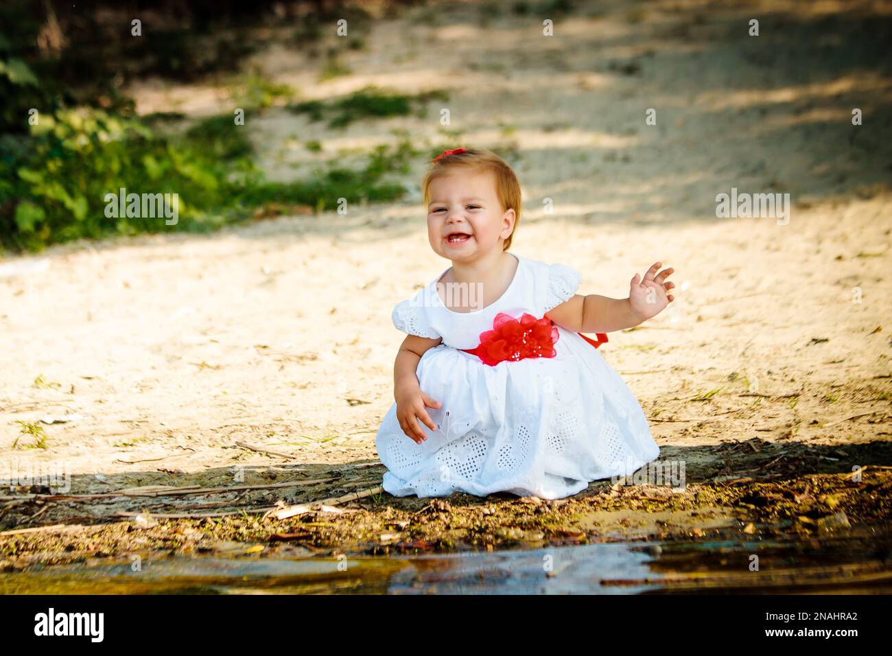 Das kleine Mädchen in einem weißen Kleid, das am Flussufer sitzt. Ein Kind, das in der Nähe des Wassers spielt. Der Junge lächelt. Das Kind grimmig und schloss seine Augen. Stockfoto