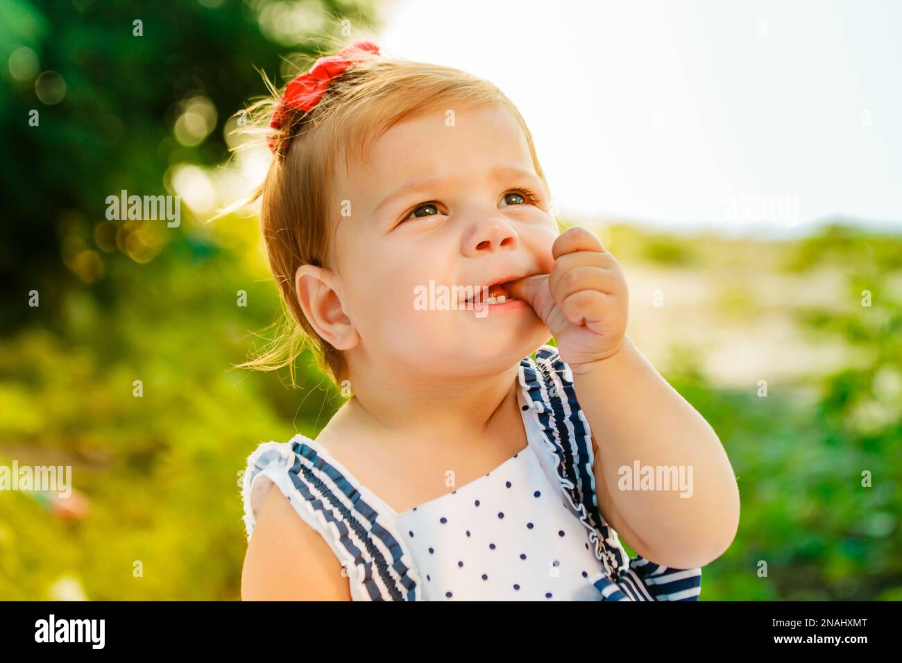 Das kleine Mädchen mit den kurzen Haaren lutscht den Finger. Das Baby schaut in den Himmel. Ein hübsches Kind, das am Ufer des Flusses sitzt, inmitten des Grüns Stockfoto