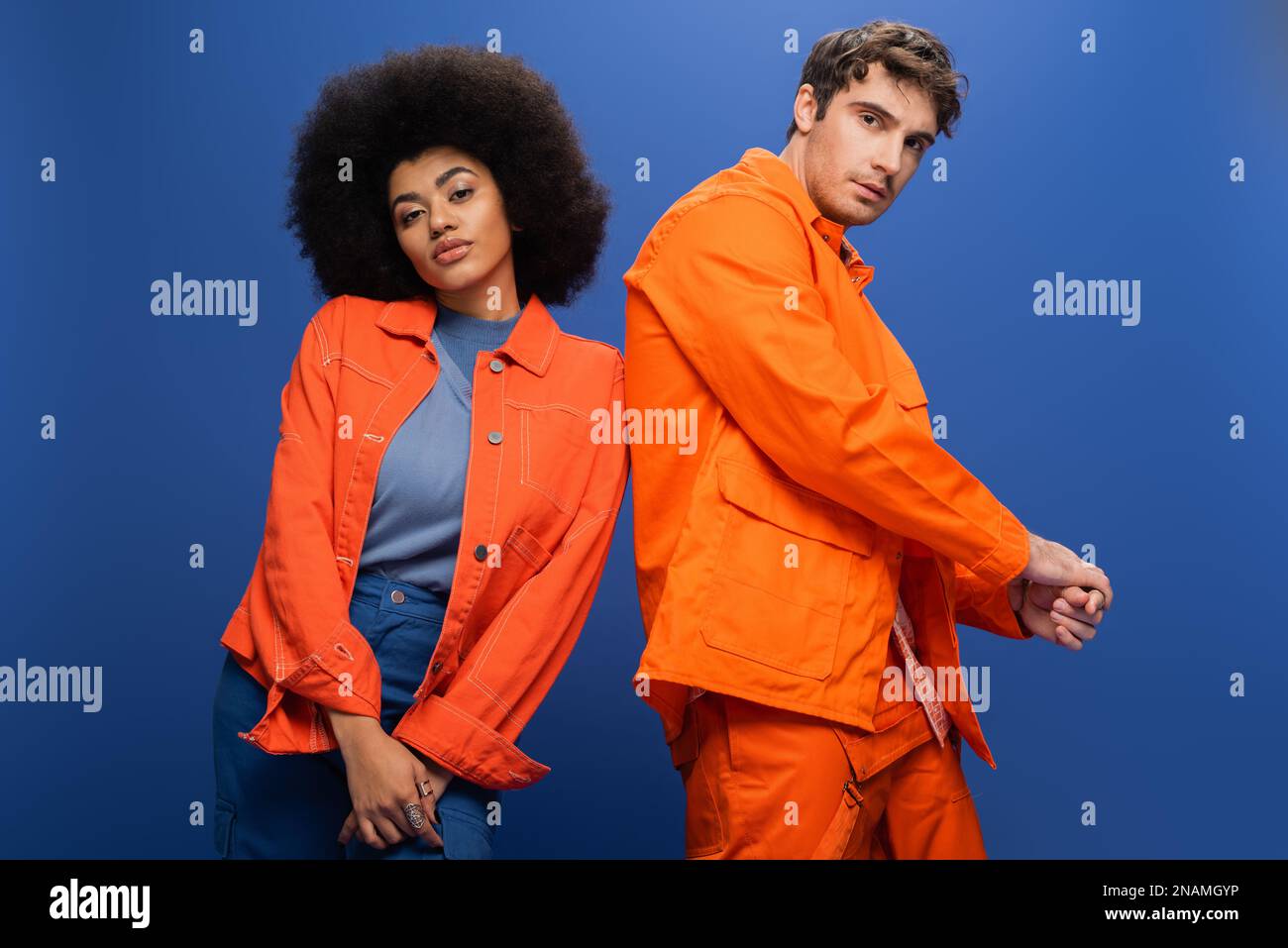 Trendiges, multiethnisches Paar in orangefarbenen Jacken, isoliert auf blauem Stockbild postiert Stockfoto