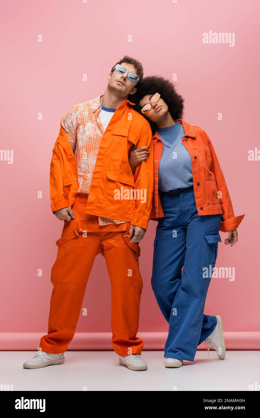 Modisches, multiethnisches Paar mit Sonnenbrille und leuchtender Kleidung, Posen auf pinkfarbenem Hintergrund, Stockbild Stockfoto