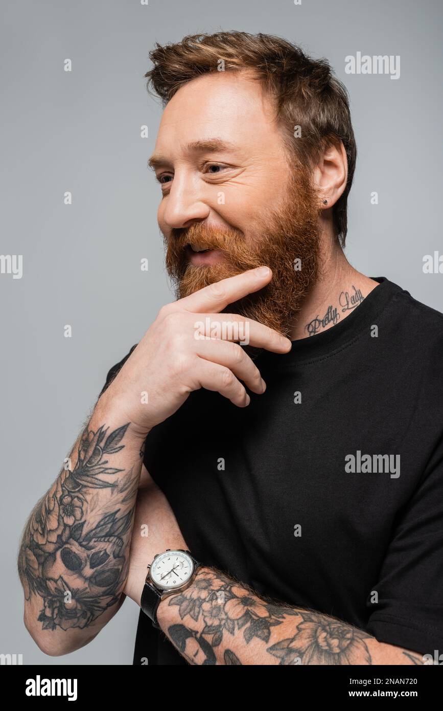 Glücklicher bärtiger Mann in schwarzem T-Shirt und Armbanduhr, der tätowierte Hand nahe dem Gesicht hält, isoliert auf grauem Stockbild Stockfoto