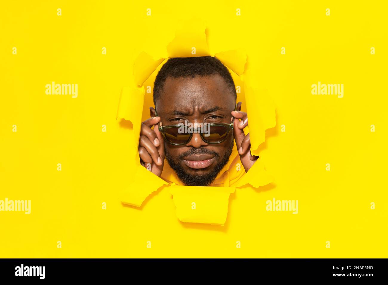 Ein afroamerikanischer Mann, der die Brille abnimmt und die Augen schielt, sieht durch ein Loch in zerrissenem gelbem Papier in die Kamera Stockfoto