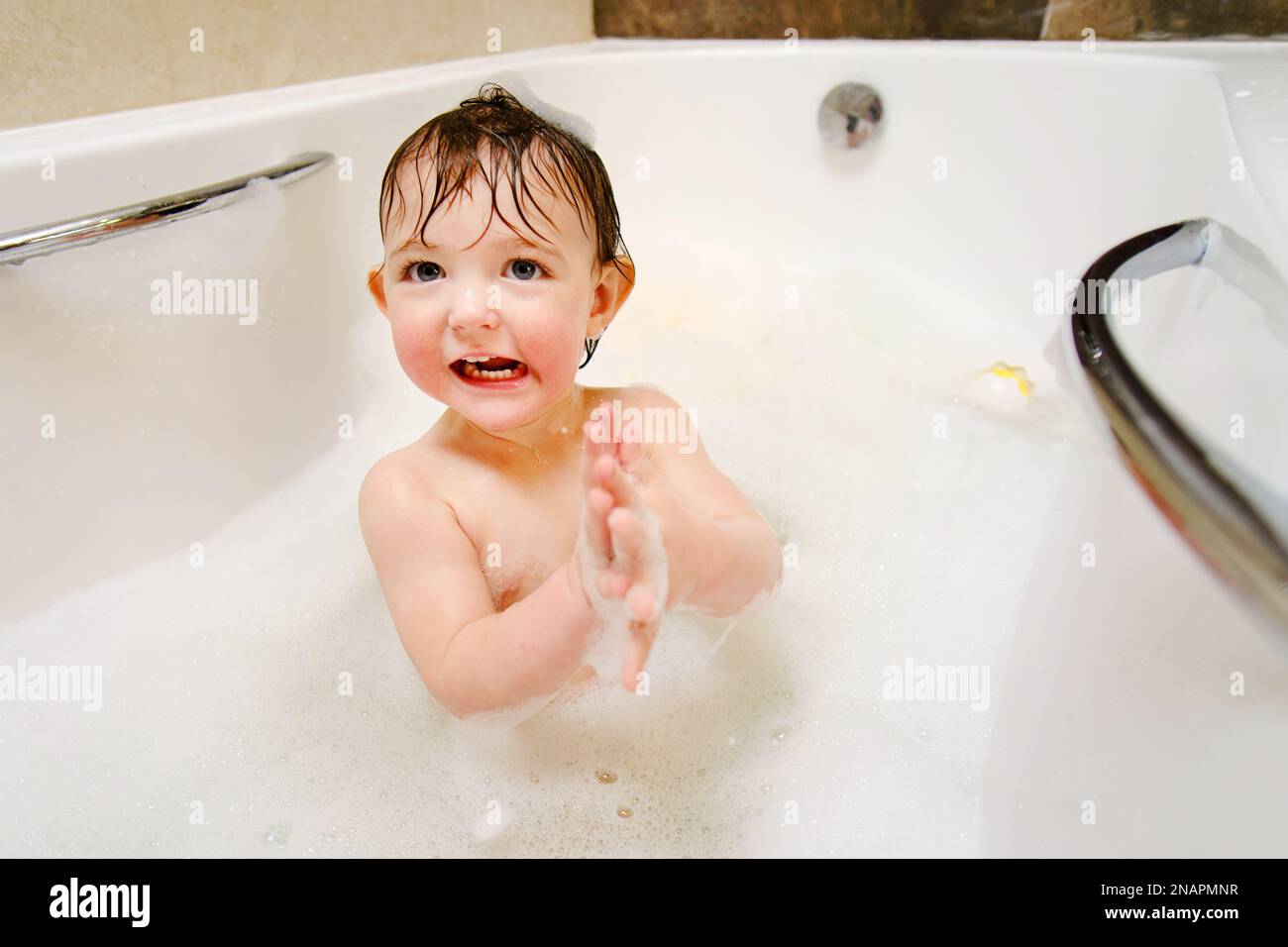 Ein glückliches Kind spielt mit Seifenblasen, während es in einer weißen Badewanne sitzt. Ein lächelnder kleiner Junge badet im Badezimmer. Ein Kind im Alter von einem Jahr Stockfoto