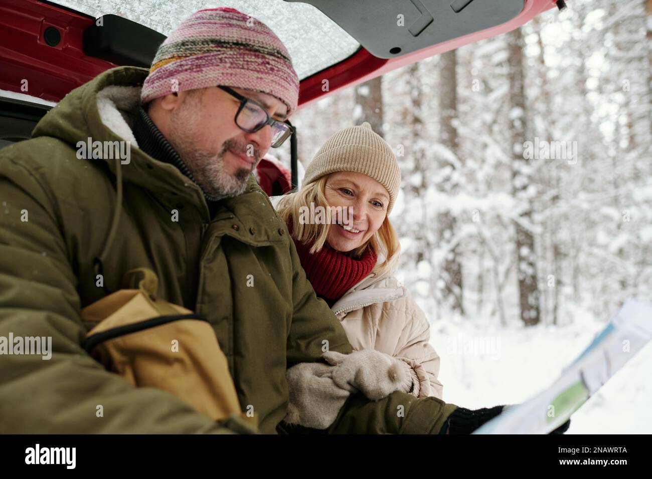 Seniorenpaar in warmer Winterbekleidung, schaute sich eine Karte an, die ein Mann in Papierform hielt, während beide auf dem Kofferraum saßen, während eine Gruppe von Bäumen mit Schnee bedeckt war Stockfoto