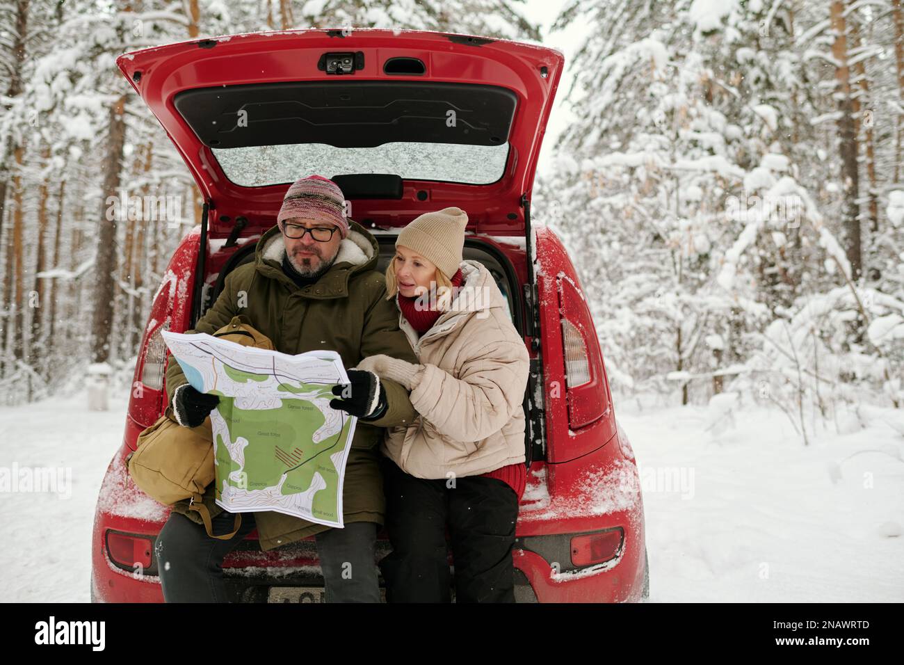 Erfahrenes Paar von Wanderer in Winterkleidung lernt Navigationskarte, während sie am Autostamm mitten im schneebedeckten Wald sitzen Stockfoto