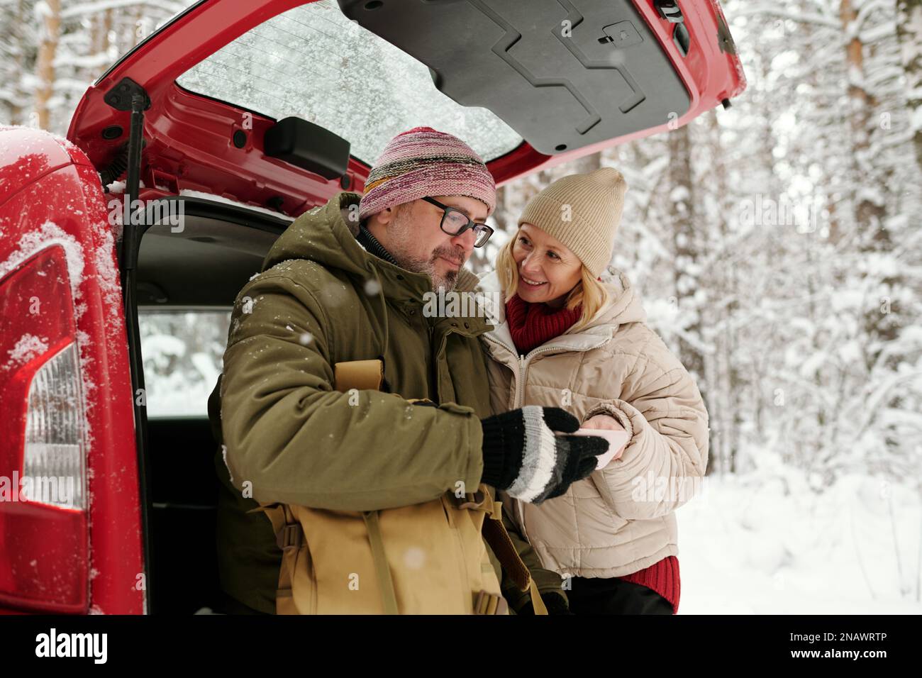 Reifer Ehemann und Ehefrau, die am Kofferraum eines Fließheckwagens stehen und entscheiden, in welche Richtung sie fahren sollen, während sie die Navigationskarte auf dem Smartphone verwenden Stockfoto