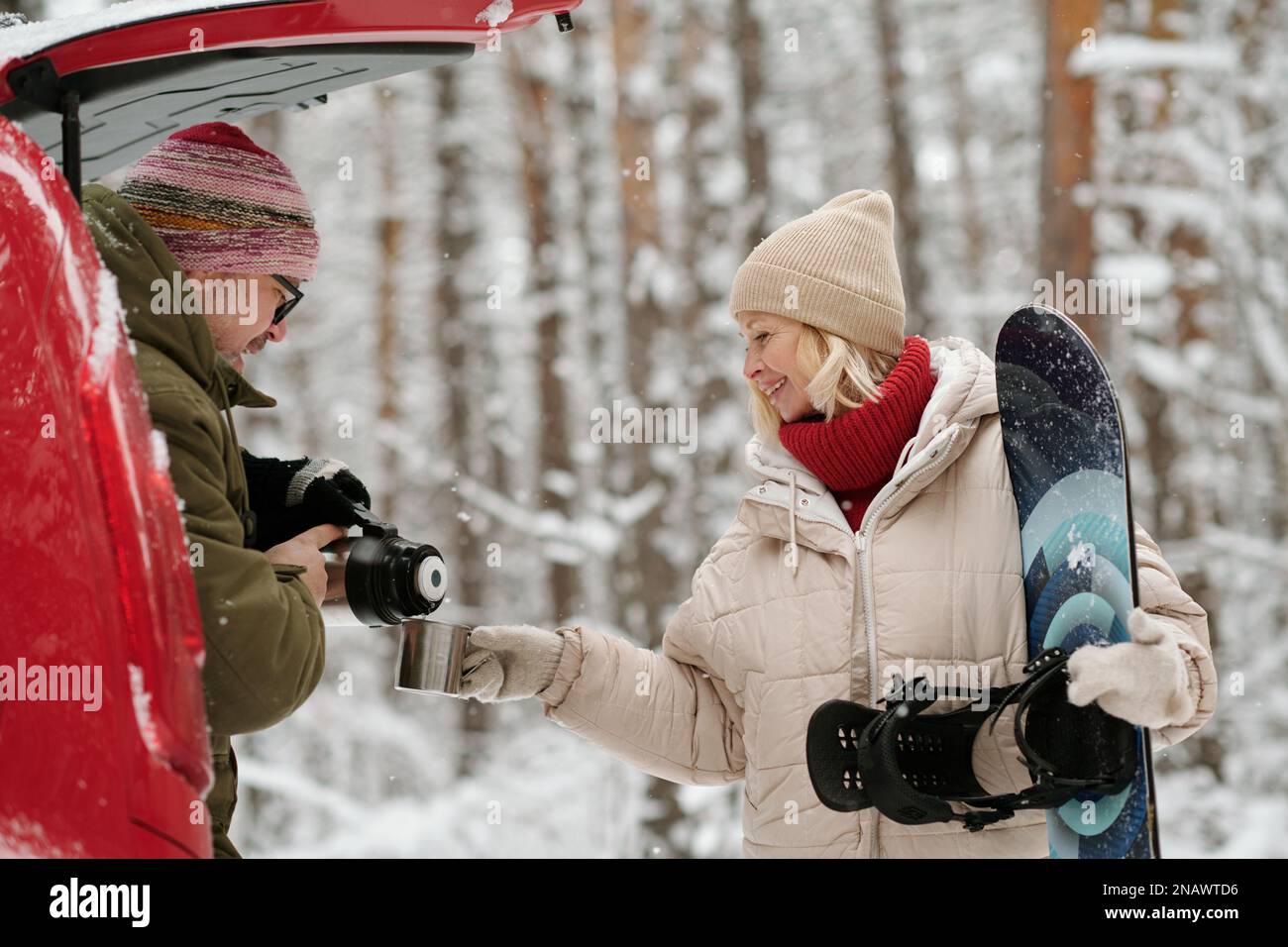 Glückliche reife Frau in Winterkleidung hält einen Metallbecher, während ihr Mann während ihrer Waldreise heißen Tee einschenkt Stockfoto