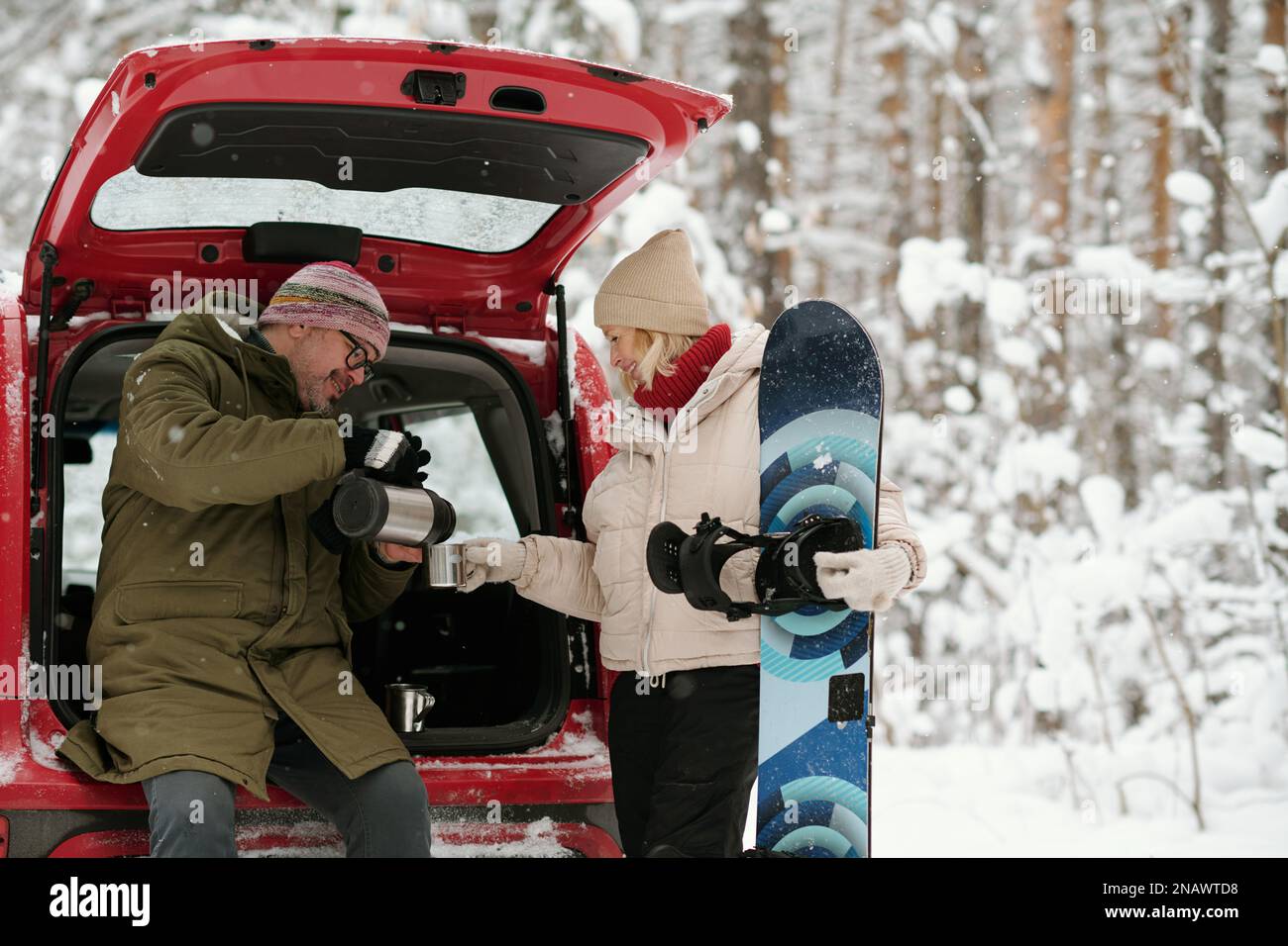 Aktiver, reifer Ehemann und Ehefrau in Winterbekleidung mit heißem Tee im Kofferraum im Wald, während eine glückliche Seniorin Skateboard hält Stockfoto
