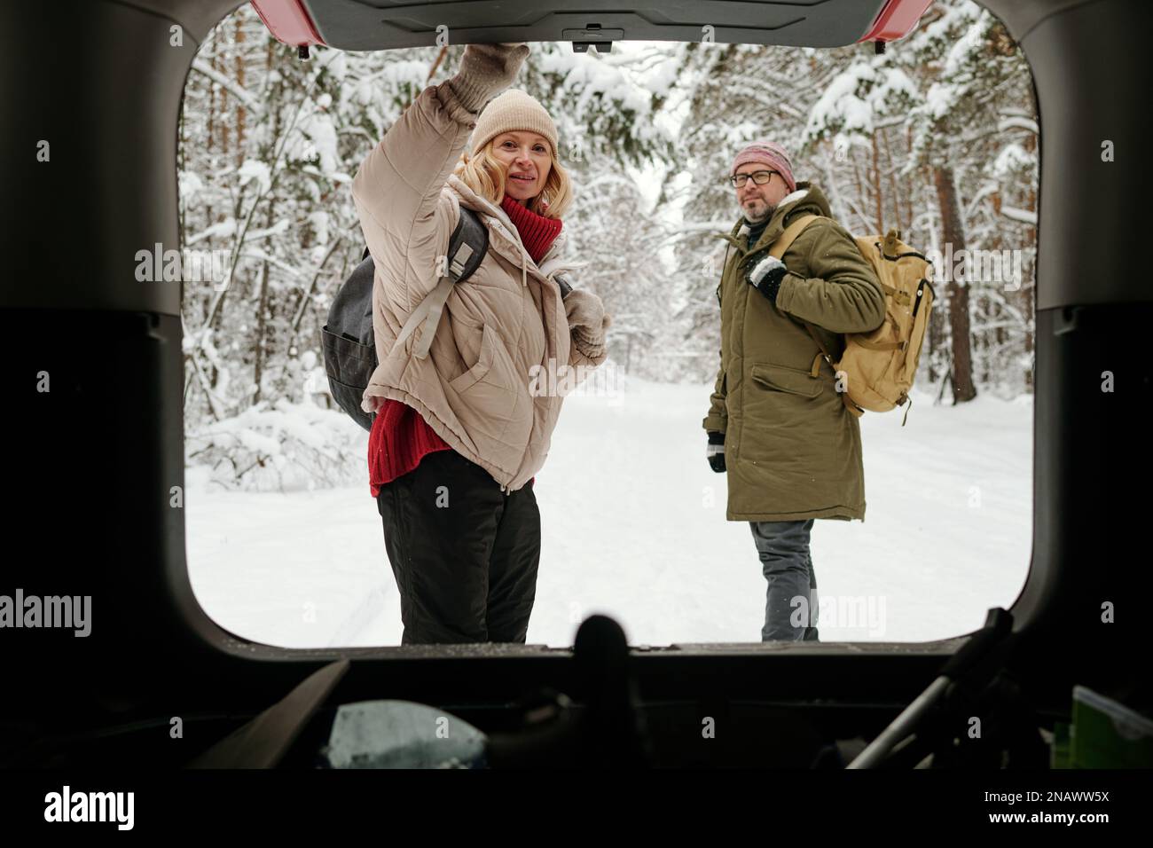Reife, blonde Frau mit dem rückentragbaren Kofferraum eines Autos, während sie im Wald auf der Straße gegen ihren Mann in Winterbekleidung stand Stockfoto