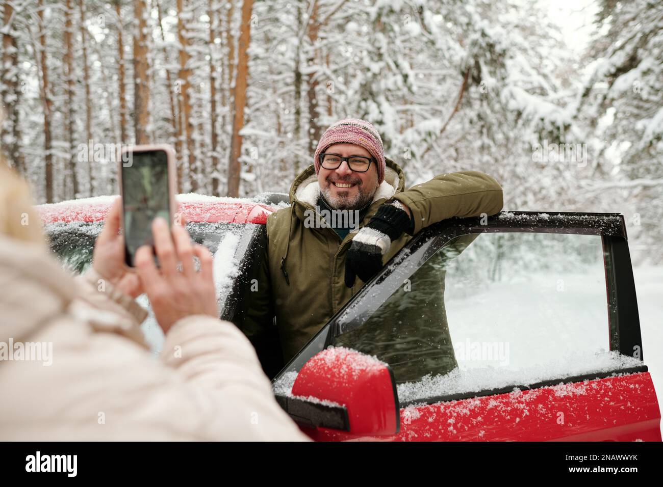 Glücklicher, reifer Mann in Brille und Winterkleidung, der im Wald an der offenen Tür des Autos stand und für seine Frau posierte, die ein Smartphone hielt Stockfoto