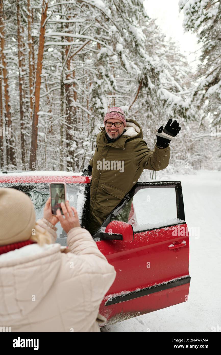 Fröhlicher, erwachsener Wanderer in Winterkleidung winkte Hand, während er in der Tür eines roten Autos stand und mit dem Smartphone für seine Frau posierte Stockfoto