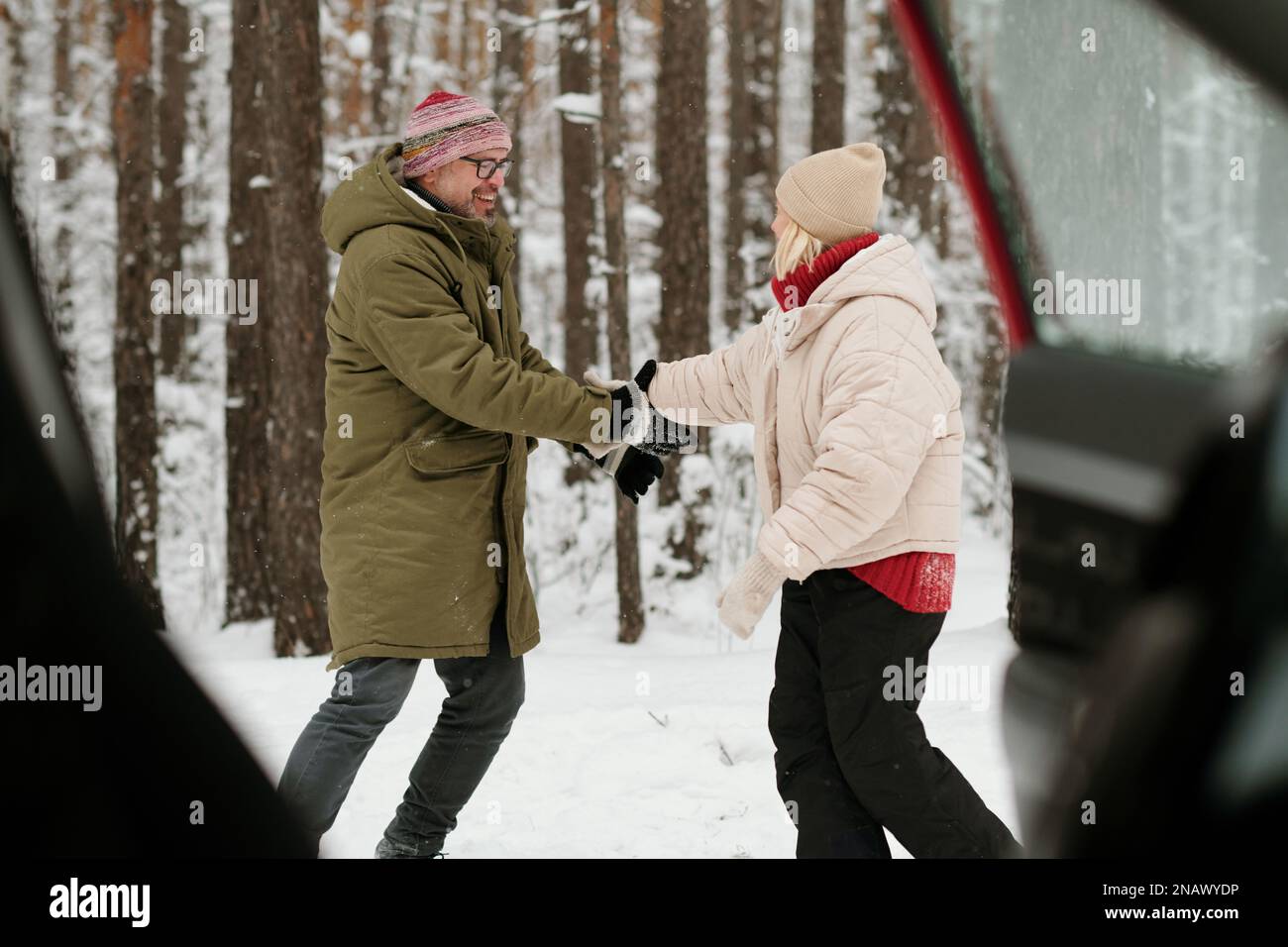 Fröhlicher Seniorenmann und -Frau in Winterbekleidung, die sich an den Händen halten und gegen Pinetrees tanzen, während sie das Winterwochenende genießen Stockfoto