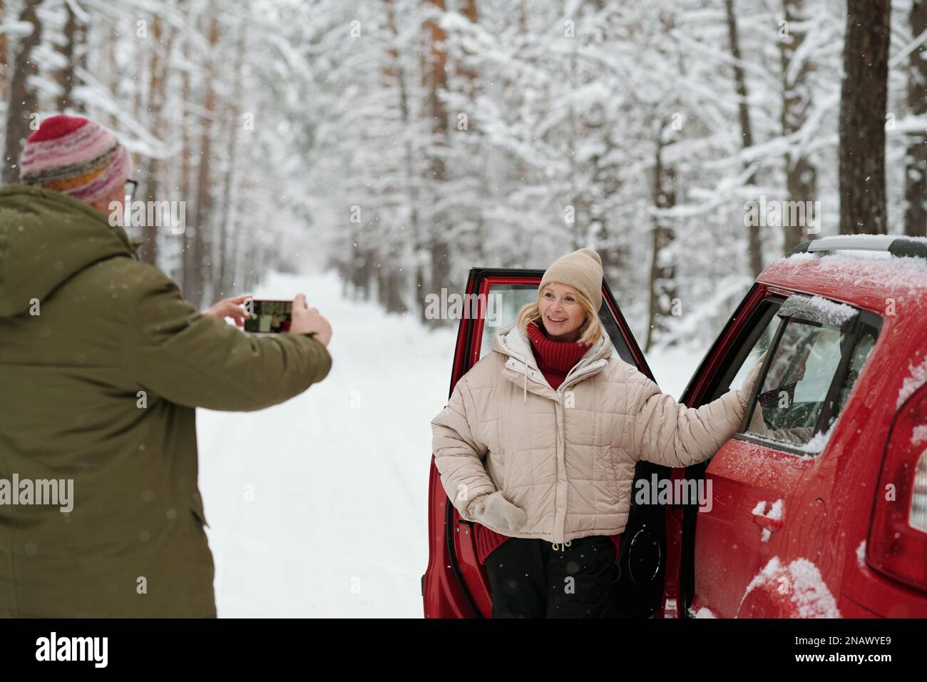 Lächelnde, reife Frau in Winterkleidung, die an der offenen Tür des Autos steht und mit einem Smartphone für ihren Mann posiert und ein Foto von ihr macht Stockfoto