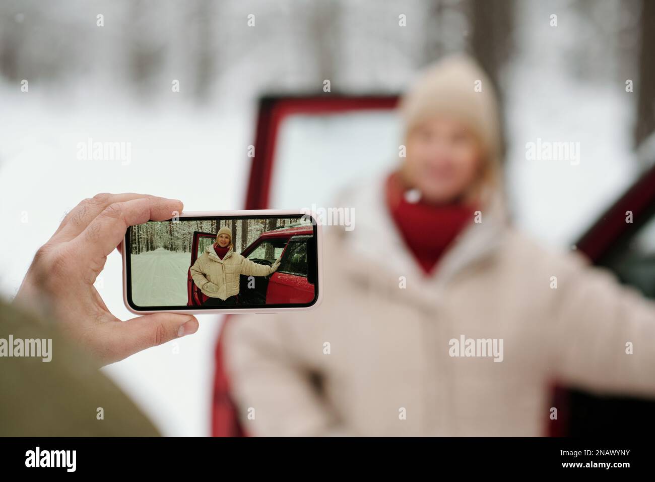 Selektiver Fokus auf die Hand eines erwachsenen Mannes, der sein Smartphone in der Hand hält, während er ein Foto seiner Frau in Winterkleidung macht, die an der offenen Tür des Autos steht Stockfoto