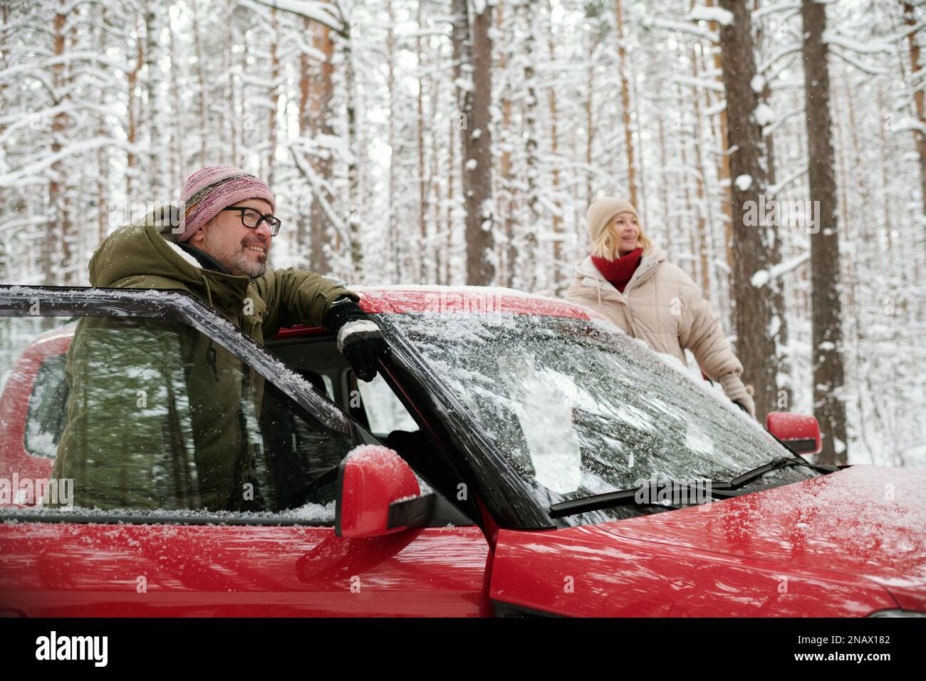 Glücklicher Seniorenmann und seine Frau in Winterbekleidung stehen in offenen Türen eines roten Autos und schauen während der Fahrt an einem verschneiten Wochenende nach vorne Stockfoto
