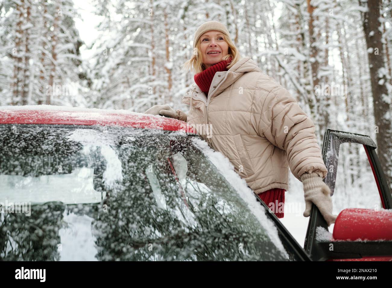 Glückliche, reife blonde Frau in warmer Winterjacke, gestrickter Beanie und Wollhandschuhe, die während der Reise an der offenen Tür des Autos stand Stockfoto