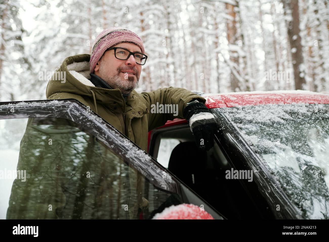 Ein lächelnder, reifer Mann in Brille und Winterkleidung steht an der Tür seines Autos und genießt Winterurlaub im schneebedeckten Wald Stockfoto