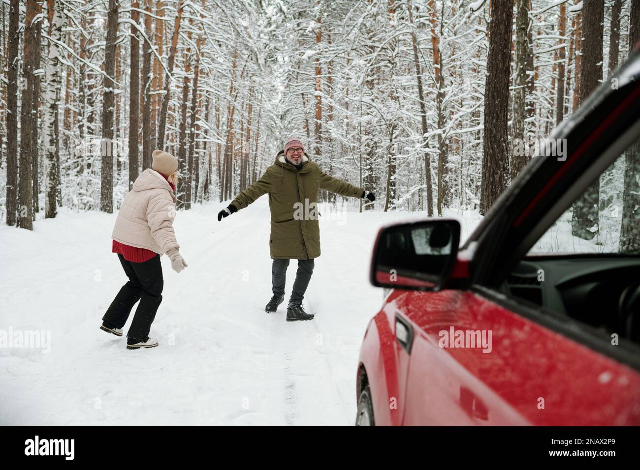 Verspieltes, reifes Paar in Winterkleidung, das auf der Straße in einem Pinetree Wald, nicht weit von seinem Auto, unterwegs ist, während des Winterurlaubs oder Wochenendes Stockfoto