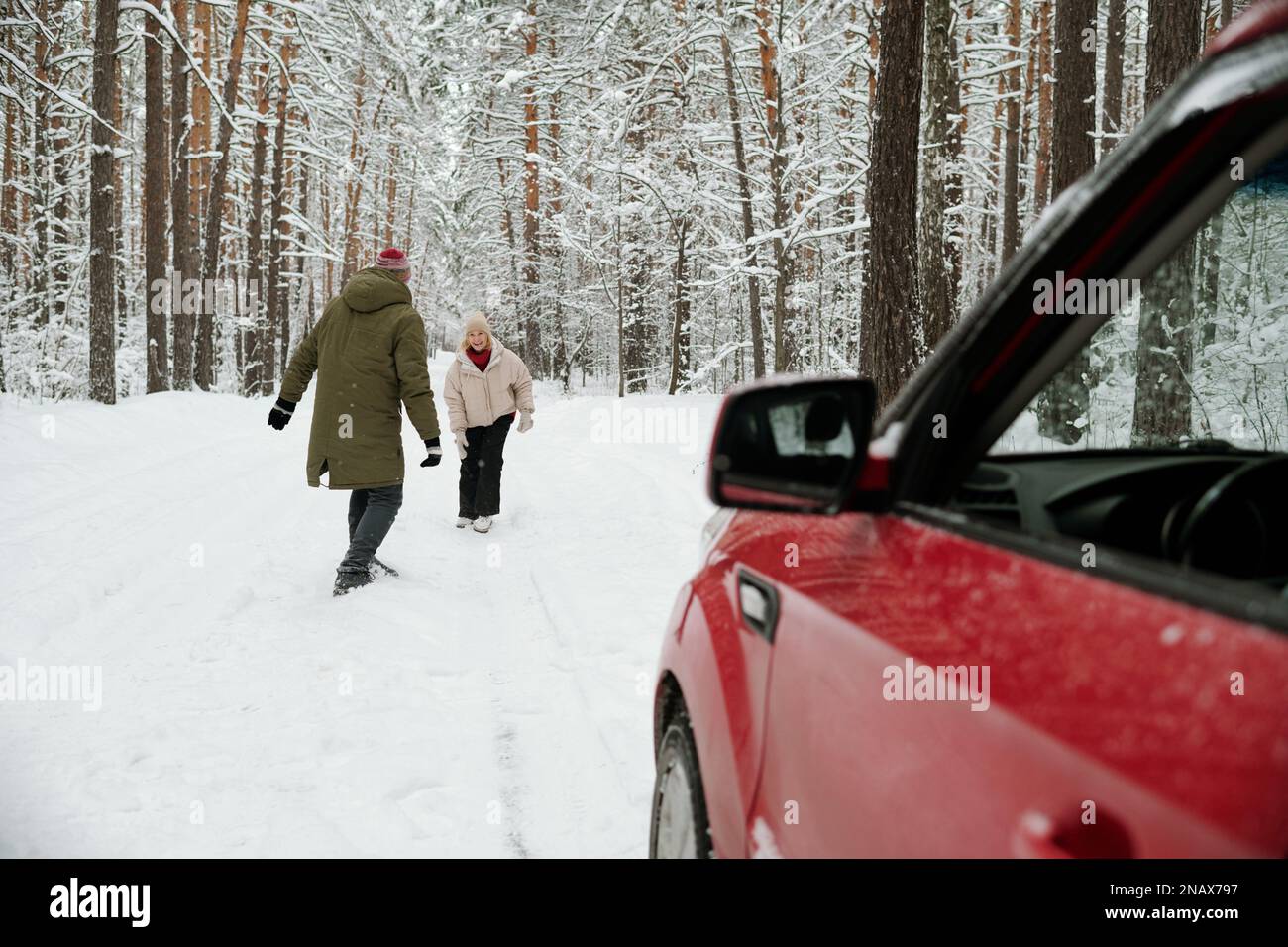 Fröhliche Seniorin in Winterkleidung, die vor ihrem Mann auf der Straße im Pinetree-Wald steht und beide am Wochenende Spaß haben Stockfoto