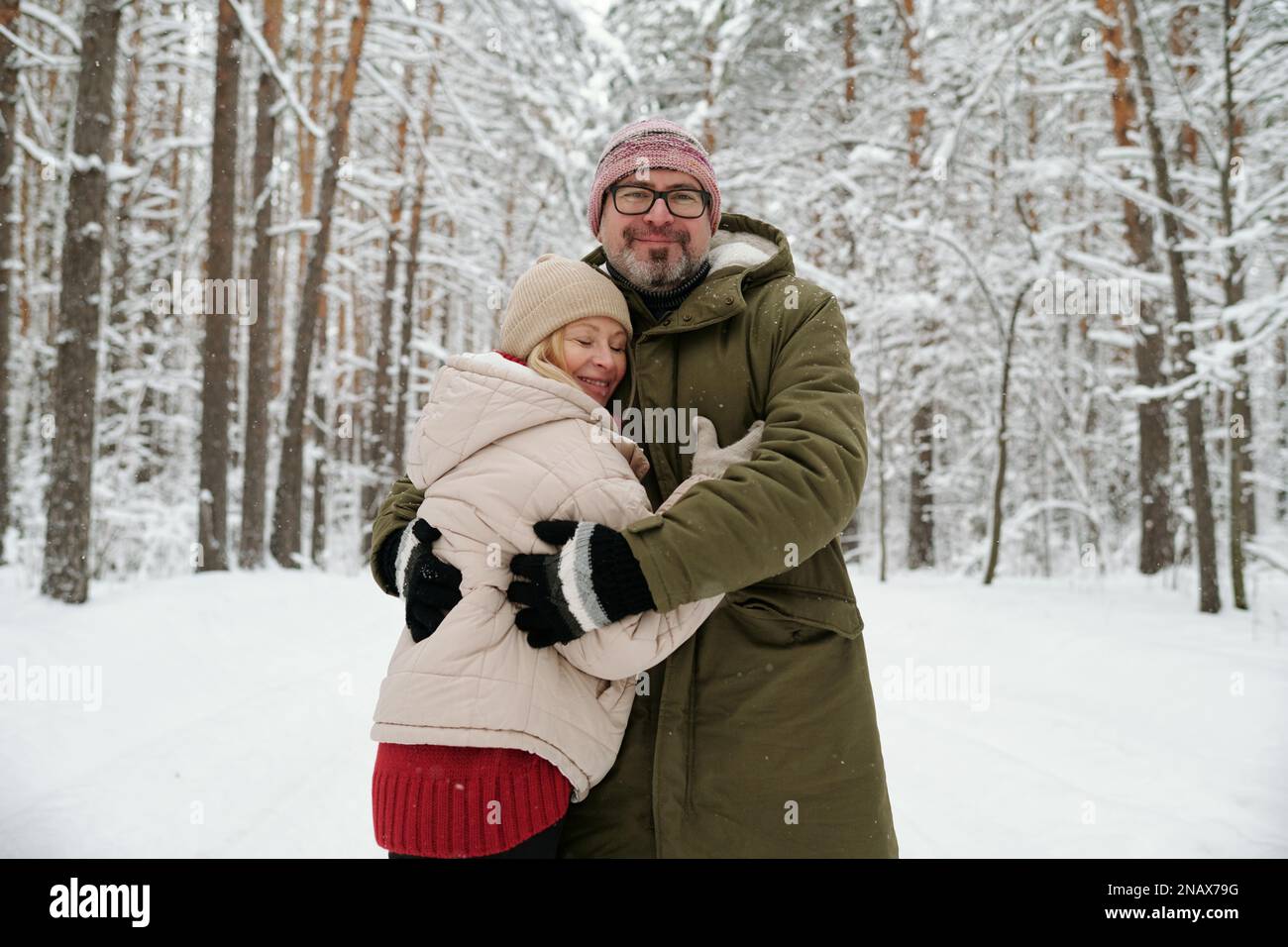 Glücklicher Senior-Mann umarmt seine Frau in warmer Winterjacke, Mütze und Handschuhen, während beide im Wald vor der Kamera stehen Stockfoto