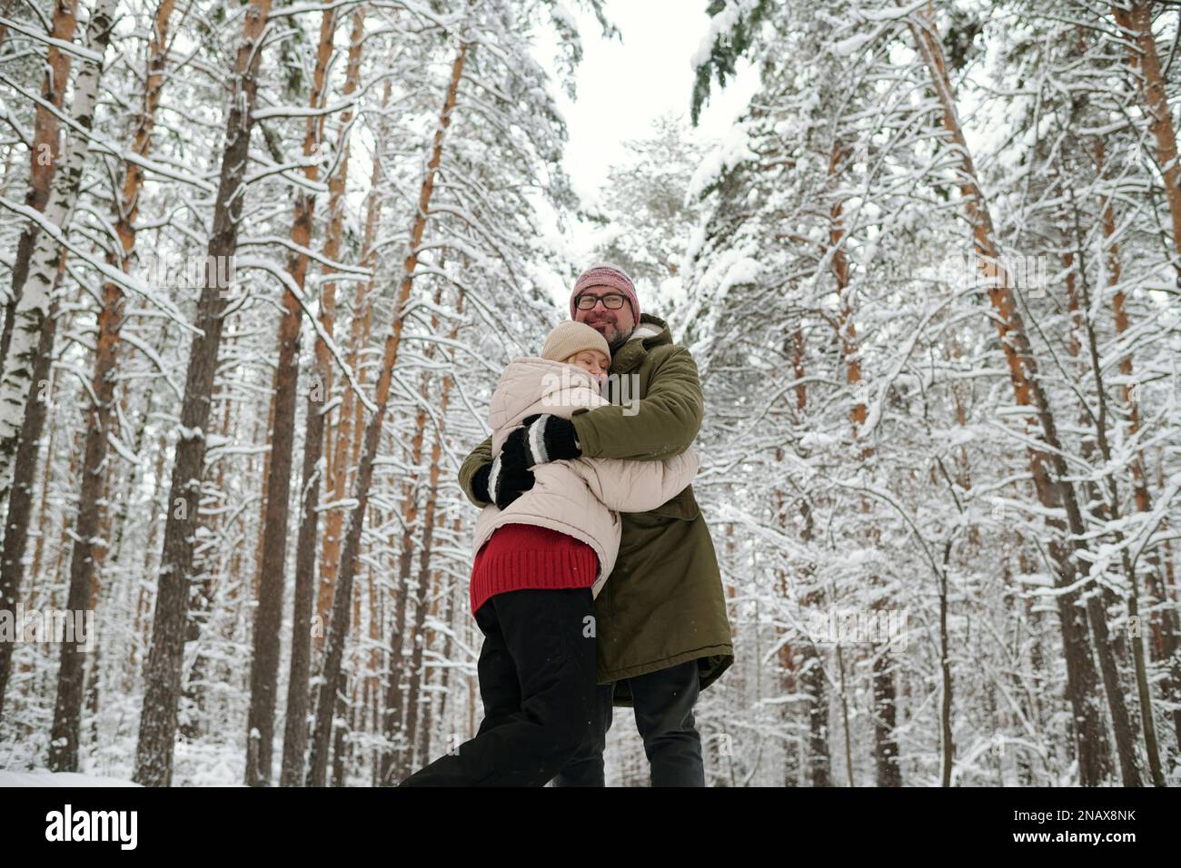 Liebevolles, reifes Paar in Winterbekleidung, das in Umarmung zwischen schneebedeckten Kiefernwäldern steht und das Winterwochenende genießt Stockfoto
